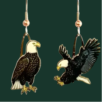 A pair of bald eagle dangle earrings displayed on a gray textured background next to a wooden ruler for scale. One earring shows a bald eagle standing, and the other shows it flying with wings spread.